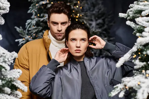 A beautiful couple embraces the holiday spirit, standing together in winter jackets amidst snowy pine trees. — Stock Photo