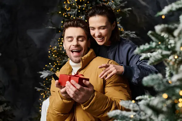 A happy couple exchanges gifts in cozy winter jackets surrounded by snow-draped pine trees. — Stock Photo