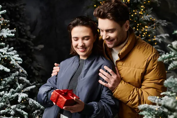 A couple in warm winter jackets smiles as they share a festive present among snow-covered pine trees. — Stock Photo