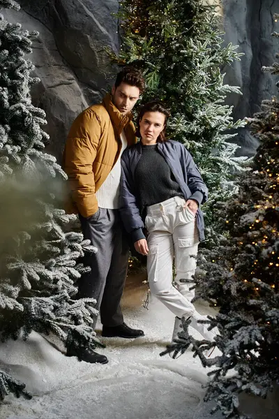 A joyful couple dressed in warm winter jackets stands among snow-covered pine trees, celebrating Christmas. — Stock Photo