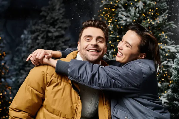 A happy couple shares a warm embrace near pine trees blanketed in snow, celebrating the Christmas spirit. — Stock Photo
