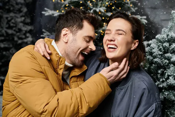 A happy couple embraces in their warm winter jackets, surrounded by snow-covered pine trees and festive spirit. — Stock Photo