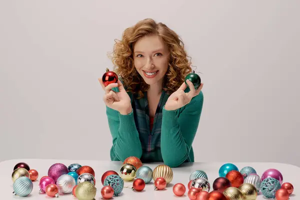 A happy young woman holds two ornaments while surrounded by festive decorations on a table. — Stock Photo