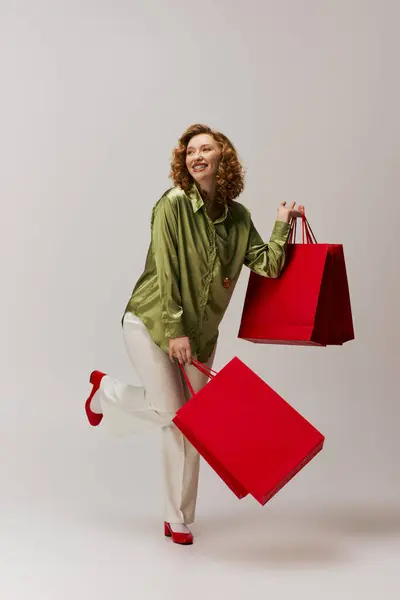 A cheerful young woman elegantly posing cheerfully while playfully holding red shopping bags, exuding joy. — Stock Photo