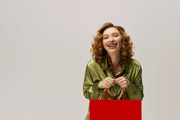 A young woman with curly hair gleefully prepares a gift with a bright red bag in a light space. — Stock Photo