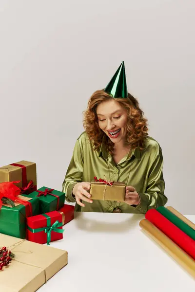 A young woman wrapped in a green shirt joyfully holds gifts while wearing a party hat. — Stock Photo