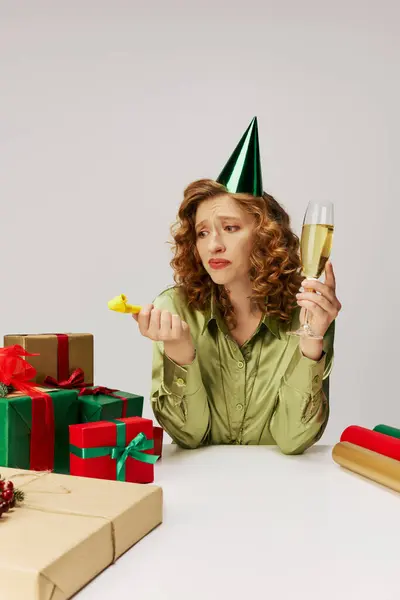 A young woman wearing a party hat sits surrounded by wrapped gifts while looking disappointed. — Stock Photo