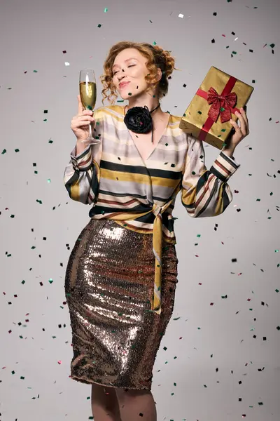 A young woman in a glamorous outfit celebrates with a drink and a beautifully wrapped gift. — Stock Photo