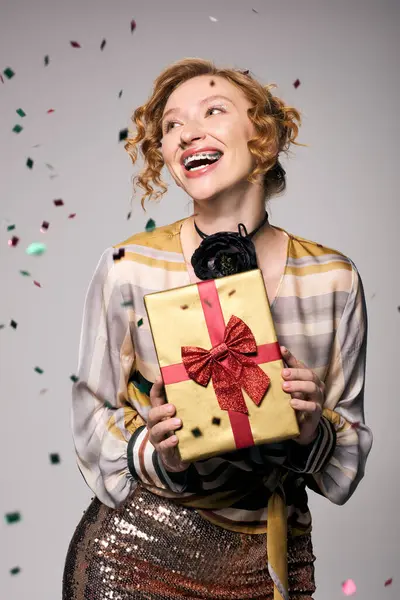 A young woman with curly hair joyfully hugs a wrapped gift amid confetti. — Stock Photo