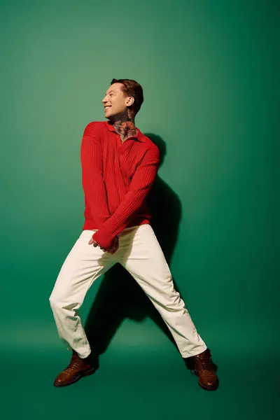 A tattooed young man joyfully poses in a bright red sweater and white pants. — Stock Photo