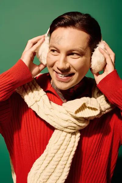 A smiling young man in a stylish sweater and earmuffs stands against a vibrant backdrop. — Stock Photo