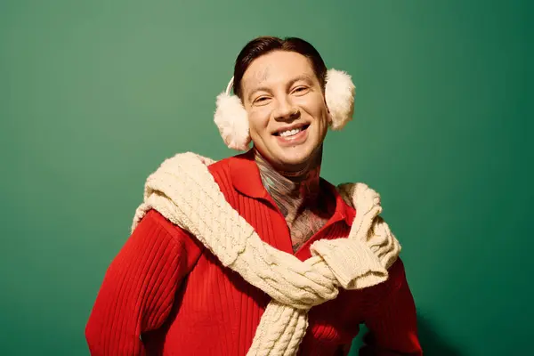 A cheerful young man with tattoos wears a vibrant red outfit and fluffy ear muffs, radiating joy. — Stock Photo