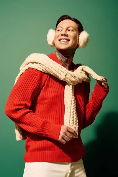 A cheerful young man with tattoos smiles while wearing a cozy red sweater and fluffy earmuffs. — Stock Photo