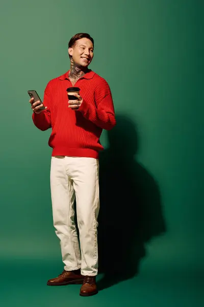 Le jeune homme vêtu d'un pull rouge tient un téléphone et une tasse de café, souriant vivement. — Photo de stock