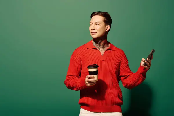 The young man with tattoos holds a coffee cup in one hand and a phone in the other while posing. — Stock Photo