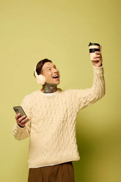 A young man sporting tattoos happily sips coffee and engages with a phone while enjoying music. — Stock Photo