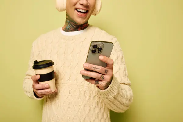 Der junge Mann lächelt, während er eine Kaffeetasse in der Hand hält und aufgeregt auf sein Handy blickt.. — Stockfoto
