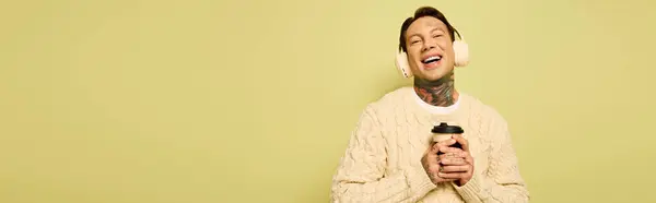 A young man with tattoos smiles broadly while holding a warm drink and wearing fluffy ear muffs. — Stock Photo