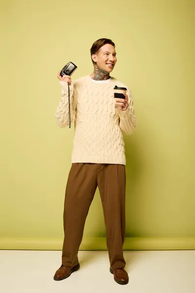 A young man with tattoos enjoys a warm coffee while holding a camera, wearing a stylish outfit. — Stock Photo