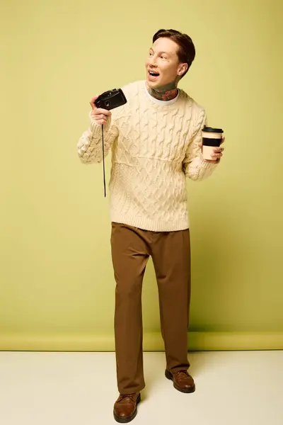A young man with tattoos joyfully balances a camera and coffee cup while posing creatively. — Stock Photo