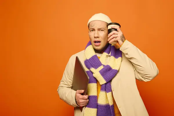 A tattooed young man in a cozy scarf looks surprised while holding a coffee cup. — Stock Photo