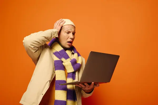 The young man, wearing a striped scarf and hat, appears shocked while using his laptop. — Stock Photo