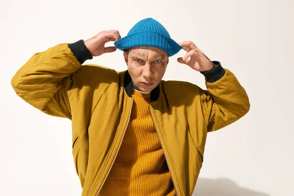A young man showcases his tattoos while adjusting a blue hat and wearing a yellow sweater. — Stock Photo