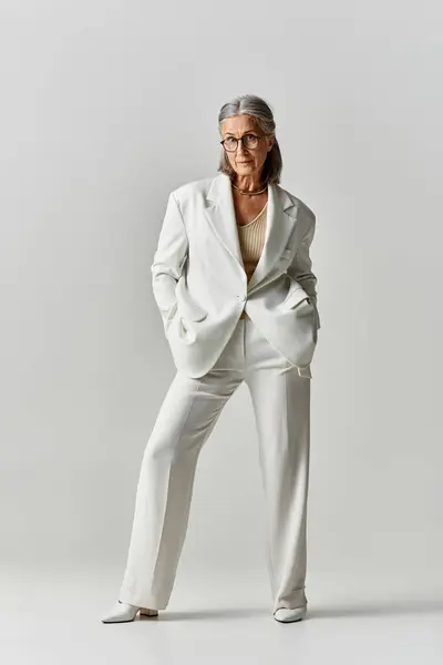 A mature woman with grey hair showcases her elegance in a chic white suit on a muted grey background. — Stock Photo