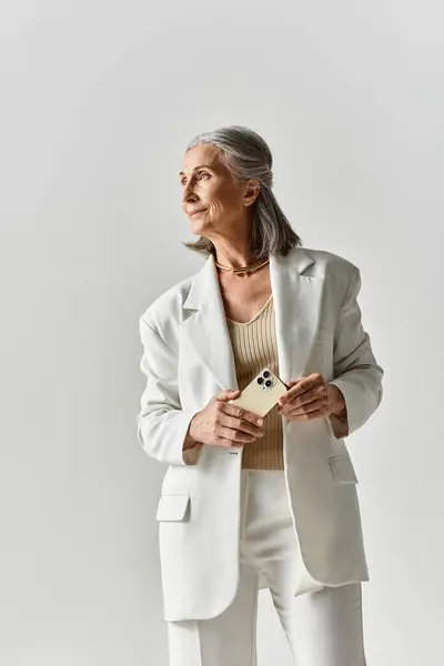 A mature woman with beautiful grey hair stands confidently in a tailored white suit, holding her phone. — стоковое фото