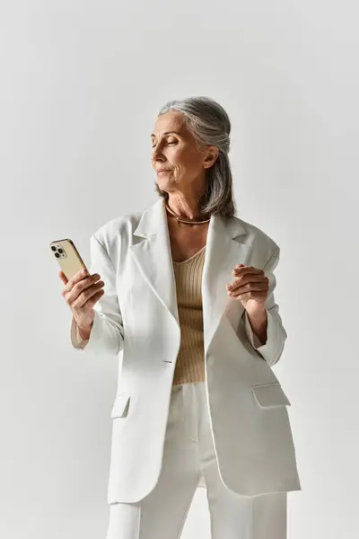 A sophisticated woman with grey hair poses gracefully in a white suit, holding a phone. — Stock Photo