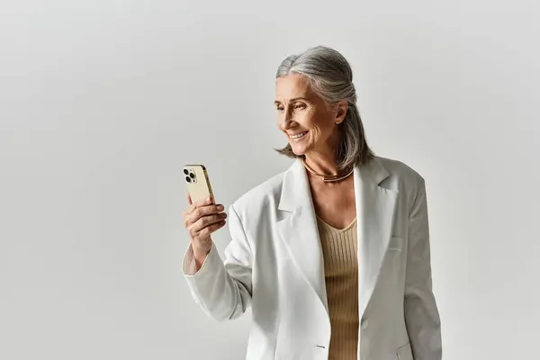 A confident, mature woman in a chic white suit smiles while looking at her smartphone, exuding elegance. — Fotografia de Stock