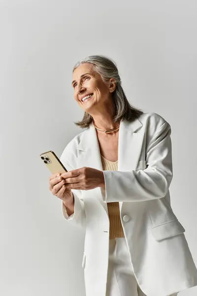 Uma mulher madura graciosa com cabelos grisalhos irradia elegância em um terno branco, sorrindo enquanto segura seu telefone. — Fotografia de Stock