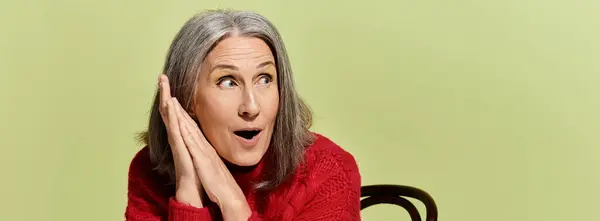Mature woman wearing a cozy red sweater expresses excitement against a bright green backdrop. — Stock Photo