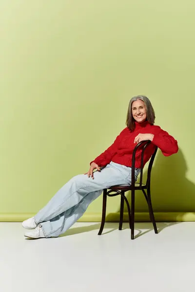 A mature woman with beautiful grey hair leans back on a chair, showcasing winter fashion in a vibrant red sweater. — Stock Photo