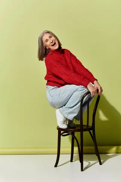 A cheerful mature woman showcases her stylish winter attire, playfully perched on a chair in a cozy setting. — Stock Photo
