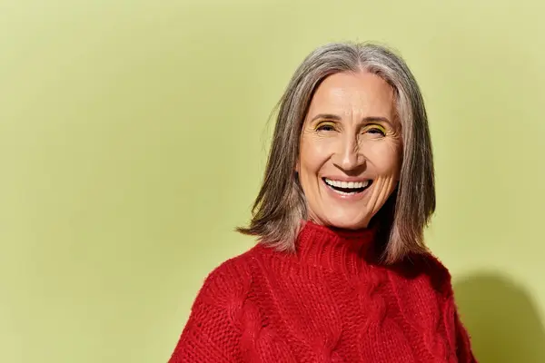 A joyful mature woman with striking grey hair exudes warmth in a cozy red sweater, set against green hues. — Stock Photo