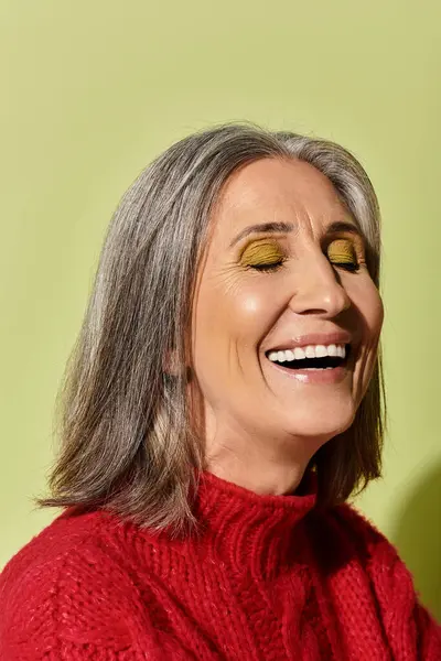 A mature woman with stunning grey hair smiles brightly, dressed in a warm red sweater against a green backdrop. — Stock Photo