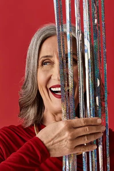 Eine fröhliche reife Frau mit grauen Haaren genießt die Weihnachts- und Neujahrsfeiern, während sie glänzende Luftschlangen in der Hand hält. — Stockfoto