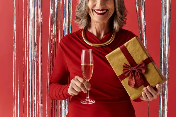 A beautiful mature woman in a festive red outfit holds a gift and a sparkling drink while celebrating. — Stock Photo