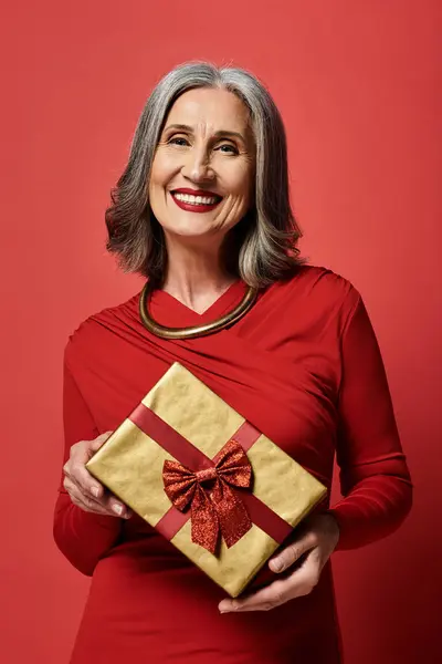 A joyful elderly woman with grey hair smiles while holding a gold Christmas gift, radiating holiday cheer. — Stock Photo