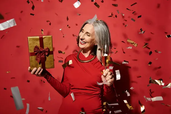 A beautiful mature woman with grey hair joyfully celebrates the holidays, holding a gift and champagne. — Stock Photo