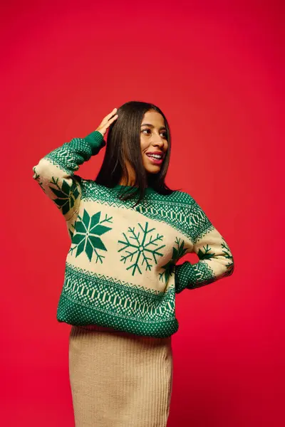 A happy young woman poses in a cozy holiday sweater against a red backdrop. — Fotografia de Stock