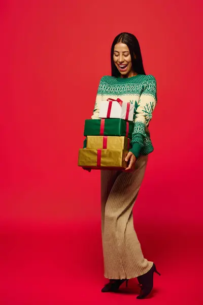 A young beautiful woman smiles while holding several wrapped gifts against a vibrant red backdrop. — Stock Photo