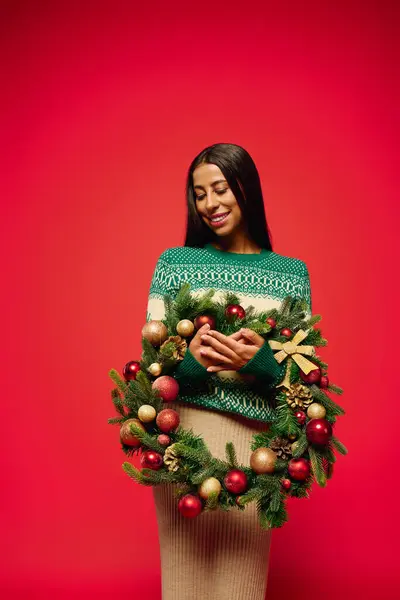 A joyful young woman beams while cradling a beautifully decorated holiday wreath. — Stock Photo