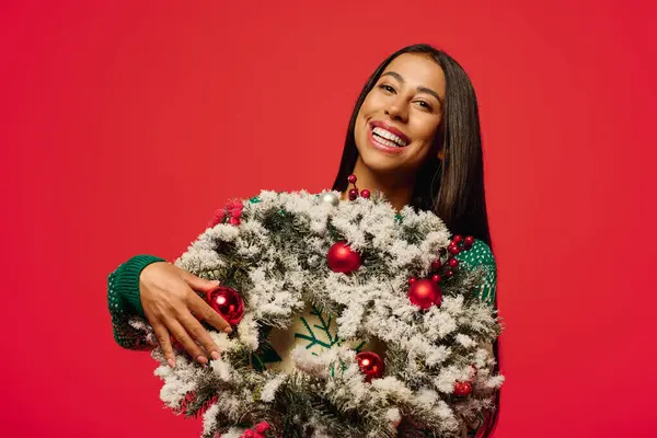 A joyful young woman smiles brightly while presenting a beautifully decorated wreath with ornaments. — Stock Photo