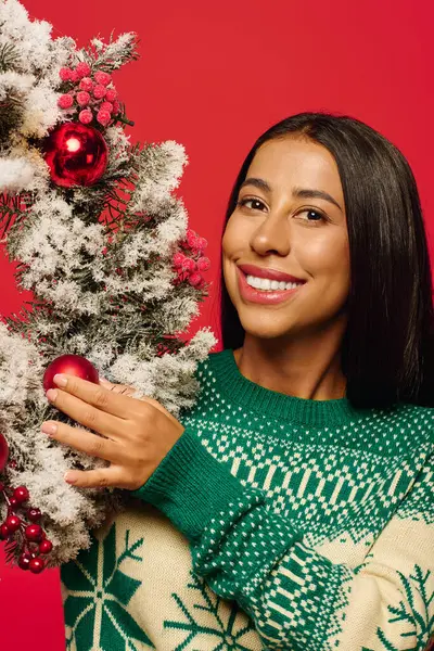 Eine fröhliche Frau im kuscheligen Pullover zeigt bunte Weihnachtsdekorationen vor hellem Hintergrund — Stockfoto