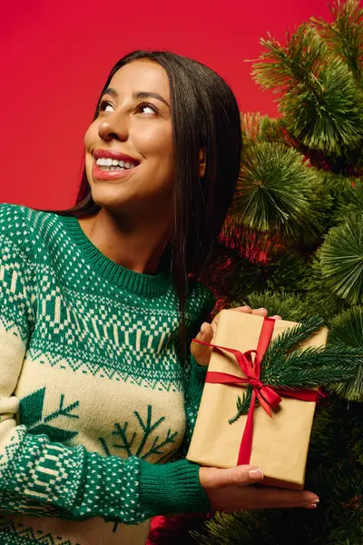 The woman, wearing a cozy holiday sweater, beams with excitement while holding a gift. — Stock Photo