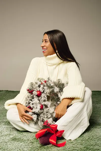 A young woman smiles while seated on soft greenery, holding a holiday wreath adorned with ornaments. — Stock Photo