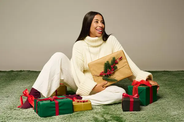 A cheerful woman in white holds a beautifully wrapped gift, surrounded by presents. — Stock Photo