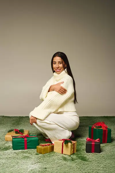 A stunning young woman in a cozy outfit kneels joyfully beside colorful wrapped gifts on soft grass. — Stock Photo